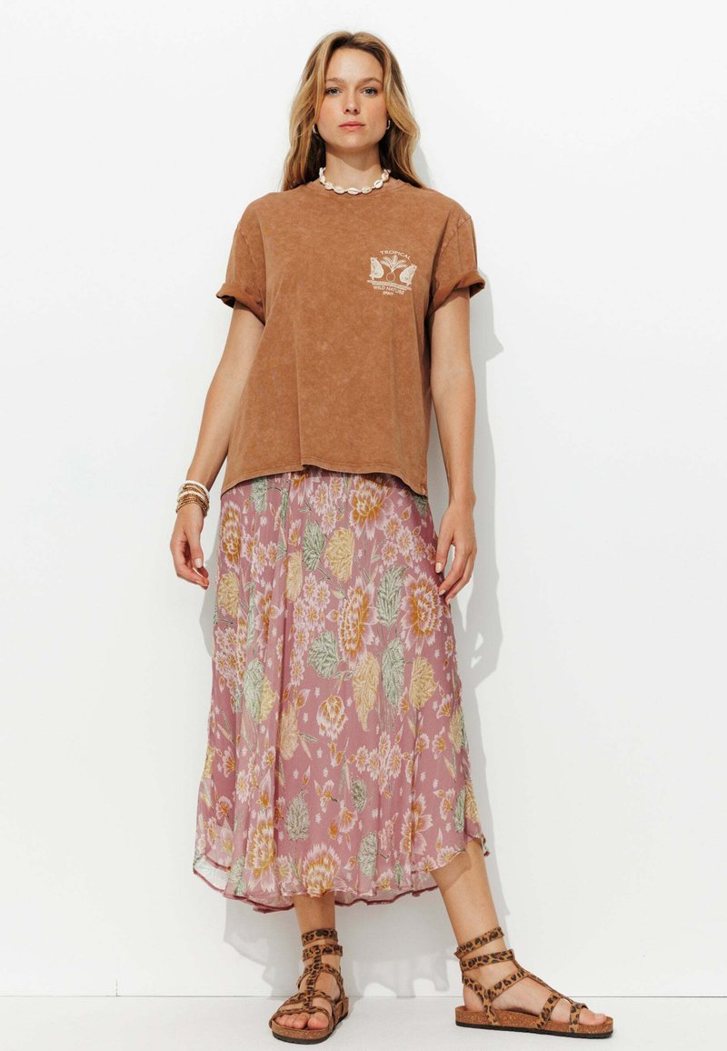 T-shirt marron à manches courtes avec un graphique, associé à une jupe midi florale rose avec des motifs jaunes et verts. Des sandales marron à lanières complètent le look.