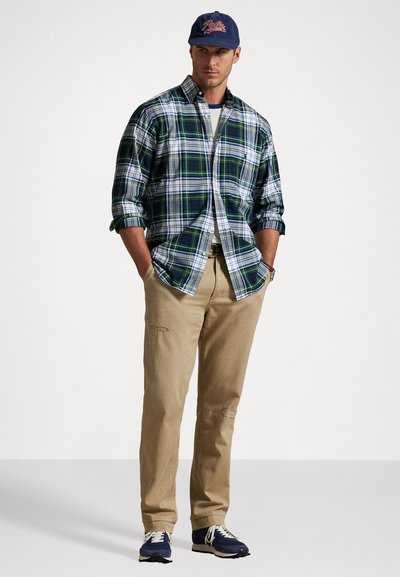 Geruit button-up shirt in groen, blauw en wit; kaki broek met subtiele details; marineblauw pet met rood logo; marineblauwe sneakers.