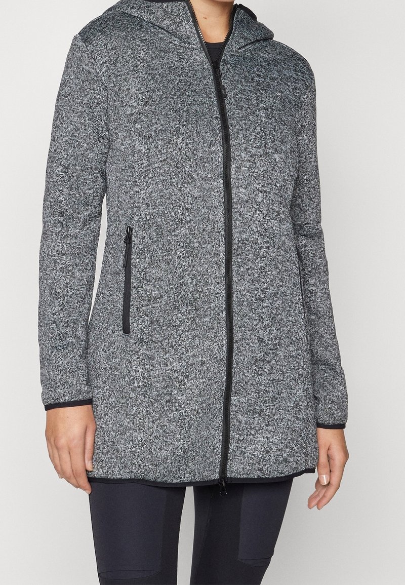 Femme portant une veste à capuche grise zippée à manches longues avec des détails de fermeture éclair et de poches noirs, associée à un pantalon noir.