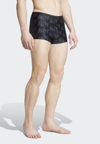 Čierne plavky s geometrickým vzorom, vyrobené z hladkého materiálu s priliehajúcim strihom. Čiastočne viditeľné nohy, nosené naboso.