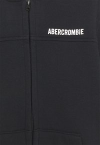 Sudadera con cremallera negra hecha de una tela suave. Presenta una cremallera frontal, puños acanalados y el logo de texto blanco "ABERCROMBIE" en el pecho.