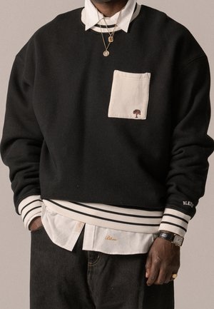 Mężczyzna ubrany w czarną warstwową bluzę z białą kieszenią i pasiastymi mankietami, noszącą pod spodem białą koszulę z kołnierzykiem, ozdobioną złotymi naszyjnikami, zegarkiem i pierścieniem.