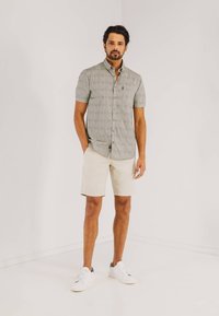 Korte mouwen, overhemd met knopen in lichtgrijs met een subtiel patroon, gecombineerd met beige shorts en witte sneakers, tegen een effen achtergrond.
