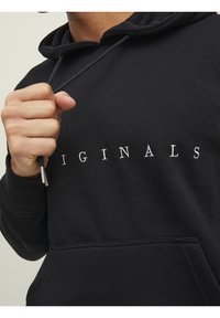 Czarna bluza z kapturem z przednią kieszenią i sznurkiem, z napisem "ORIGINALS" na piersi w białych literach. Gładka tekstura tkaniny.