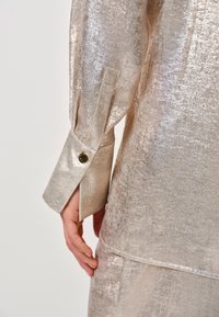 Metallic gold blouse with long sleeves. Features a buttoned cuff and textured surface, reflecting light with a soft sheen.
