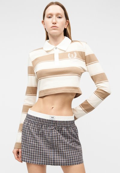 Νεαρή γυναίκα στέκεται με το ένα χέρι στη μέση, φορώντας μπεζ και λευκό ριγέ κοντό πόλο μπλουζάκι και μπλε μίνι φούστα με καρό σχέδιο, σε λευκό φόντο.