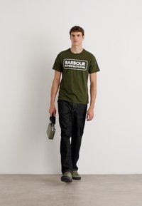Olivově zelené bavlněné tričko s logem "BARBOUR INTERNATIONAL", ve spojení s tmavými kalhotami a šedými botami. Model drží šedou tašku.