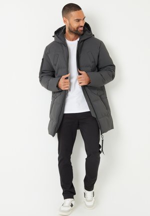 Tmavě šedá prošívaná bunda s kapucí, opatřená zapínáním na zip, dvěma předními kapsami a texturovaným povrchem, nošená s bílou košilí a černými kalhotami.
