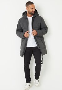 Giacca imbottita grigio scuro con cappuccio, dotata di chiusura con zip, due tasche frontali e finitura testurizzata, indossata con una camicia bianca e pantaloni neri.