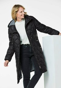 Schwarzer, gesteppter langer Mantel mit Kapuze, mit einem Reißverschluss vorne, zwei Seitentaschen und einem schimmernden Finish, getragen über einem hellen Oberteil und dunklen Jeans.