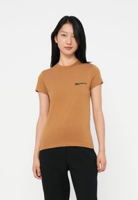 Jonge vrouw met steil zwart haar, gekleed in een aansluitend bruin T-shirt met korte mouwen en zwarte logo tekst, en zwarte broek, staande tegen een witte achtergrond.