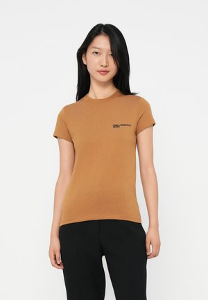 Jonge vrouw met steil zwart haar, gekleed in een aansluitend bruin T-shirt met korte mouwen en zwarte logo tekst, en zwarte broek, staande tegen een witte achtergrond.