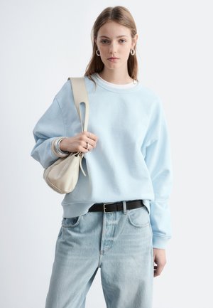 Ung kvinde iført lyseblå sweatshirt og jeans holder en beige skuldertaske og står mod en ensfarvet hvid baggrund.
