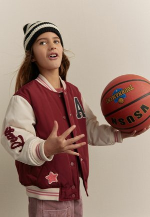 Rød og cremefarvet varsity-jakke med broderede bogstaver og stjernedetaljer. Iført en sort og hvidstribet beanie og holder en basketball.