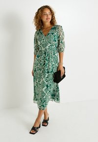 Robe midi verte à motifs avec un décolleté en V, des manches trois-quarts et une taille cintrée, associée à des sandales à talons blocs noires et à une pochette.
