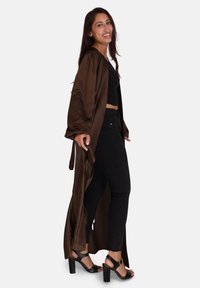 Bruine satijnen lange cardigan met losse mouwen, gedragen over een zwarte crop top en zwarte jeans. Heeft een open voorkant en strikjes in de taille. Zwarte haksandalen.