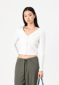 Cardigan blanc à côtes avec un col en V, fermeture à boutons et manches longues, associé à un pantalon taille haute vert olive et un sac bleu.