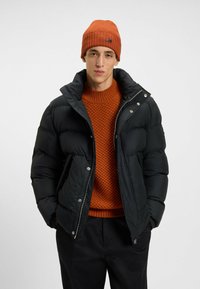 Zwarte pufferjack met een hoge kraag, drukknoopsluiting, gedragen over een oranje gebreide trui. Model draagt een bijpassende oranje beanie.