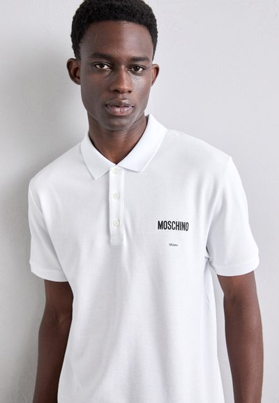Valkoinen polo-paita puuvillakankaasta, jossa on klassinen kaulus, musta Moschino-logo ja "Milano"-teksti, nappisuljenta ja lyhyet hihat.