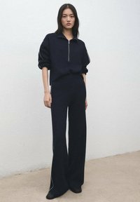 Tmavě modrá pletená tepláková souprava se zipovým límcem, volným střihem, dlouhými rukávy a proužky po stranách; doplněná jednoduchou černou obuví.