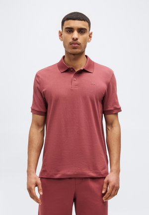 Giovane uomo che indossa una polo e pantaloni coordinati color rosa antico, in piedi con le braccia rilassate, rivolto verso l'obiettivo su uno sfondo uniforme.