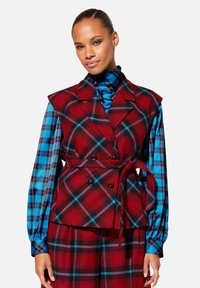 Rood geruite vest met een wikkeldesign, voorzien van een kraag en dubbelknopsluiting, gelaagd over een blauw-zwart geblokt shirt.