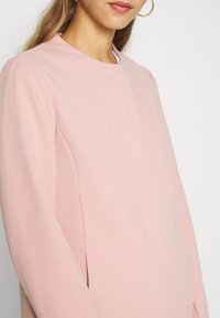 Top ajustado de color rosa claro con cuello alto y mangas largas. Textura suave y detalles de costura sutiles. Acento de pendientes de aro dorados.