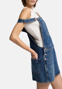 La salopette in denim presenta un lavaggio blu, spalline regolabili, una grande tasca frontale e dettagli con bottoni sul bavaglino e sui lati. È visibile un tessuto dalla texture.