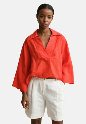 Vrouw draagt een losse rode V-hals blouse met lange mouwen, gestopt in een witte, hooggetailleerde short, met handen in de zakken, tegen een effen achtergrond.