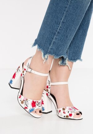 Sandales à talon bloc blanches avec broderie florale rouge, bleue et brune portées avec un jean bleu à ourlet effiloché.