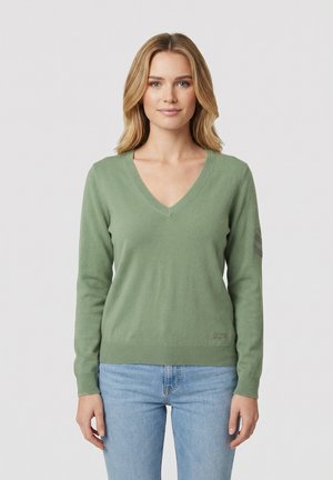 Femme aux cheveux blonds portant un pull vert clair à col en V et un jean bleu clair, debout sur un fond blanc uni.