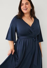 Marineblauwe wikkeljurk van zachte stof met korte, uitlopende mouwen, een diepe V-hals en een gestrikt taille, met geplooide details in de rok.