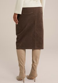 Fustă midi din cord din bumbac maro, cu două buzunare la spate, purtată cu cizme bej cu toc înalt, care ajung până la genunchi. Prezintă o textură netedă și un design ajustat.