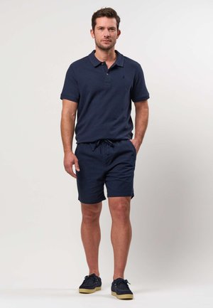 Mand iført navy polo-shirt, matchende shorts og mørke sneakers med gummisåler mod en ensfarvet lys baggrund.