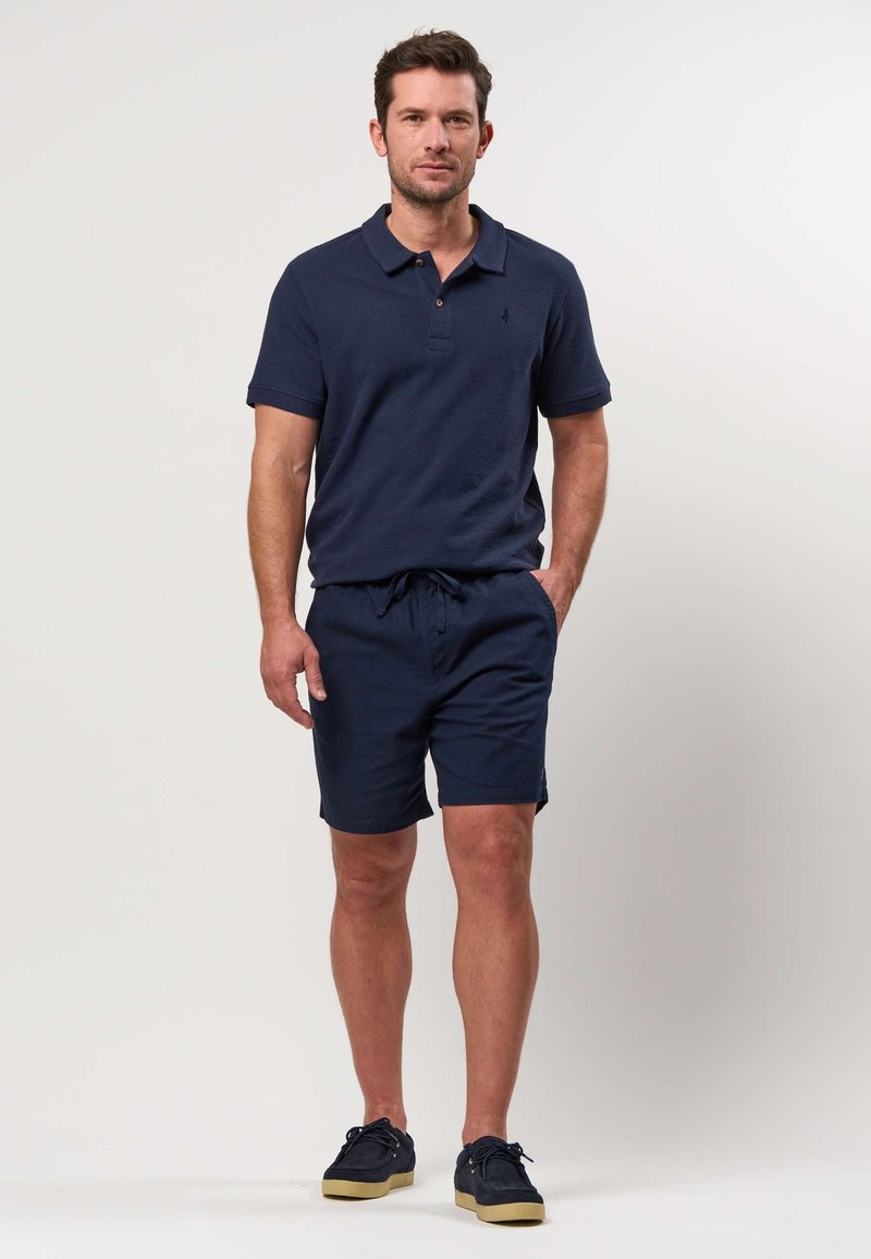 Homme debout portant un polo bleu marine, un short assorti et des baskets foncées à semelles en gomme devant un fond clair uni.