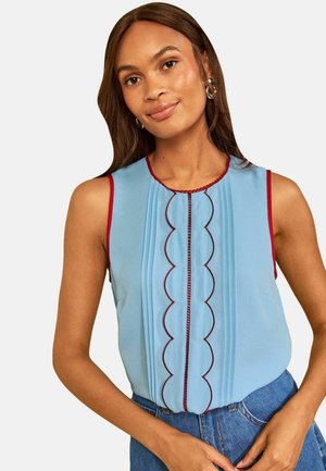 Jeune femme aux longs cheveux bruns porte un chemisier bleu clair sans manches avec une bordure rouge en festons et un jean bleu taille haute, souriant doucement.