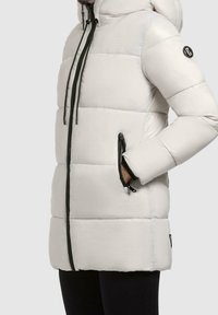 Chaqueta blanca acolchada con capucha, que presenta cremalleras negras y un logo. Tiene bolsillos laterales, un acolchado geométrico y una textura suave y brillante.