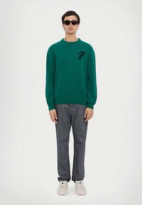 Zaļš adīts džemperis ar apaļu kaklu, uz krūtīm melns 7. numura logotips, kombinēts ar pelēkajām džinsu biksēm un baltām sporta kurpēm.