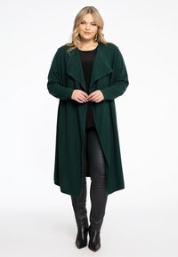 Cardigan vert long en tissu doux, avec un devant ouvert drapé et des manches larges, associé à un haut et un pantalon noirs. Bottes pointues.