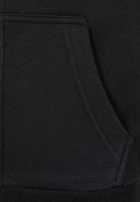 Nærbilde av svart stoff med kengurulomme, synlige sømmer og ribbestrikket kant på et plagg.