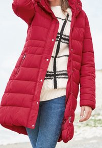 Abrigo rojo acolchado con capucha, que presenta un diseño acolchado y cierres a presión, llevado sobre un suéter crema con patrones de cuadros negros y jeans azules.