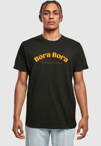 Melnā kokvilnas T-krekls ar uzrakstu "Bora Bora" bold dzeltenā rakstībā un "FRENCH POLYNESIA" zemāk mazākā sarkanā tekstā. Standarta izmērs.