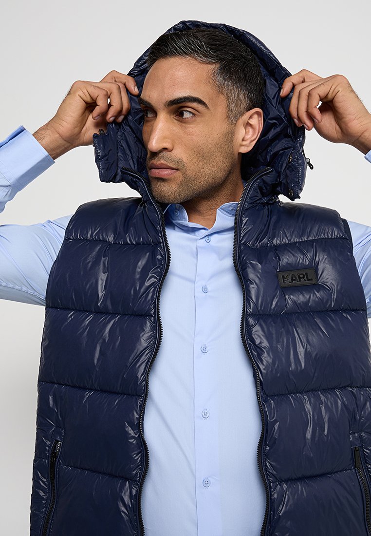 Gilet matelassé bleu marine avec une texture brillante, fermeture éclair et deux poches avant. Dispose d'une capuche amovible et d'un patch logo noir.