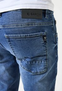 Jeans de mezclilla en azul medio, con un sutil efecto de desgaste, múltiples bolsillos y una etiqueta de cuero negro con el texto "GARCIA" en la cintura.