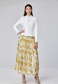 Veste blanche texturée avec des boutons associée à une jupe longue à motifs dorés et fleuris. Modèle portant des talons blancs à lanières.