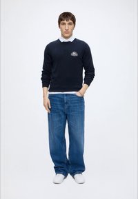 Jeune homme debout portant un pull bleu marine sur une chemise blanche à col, un jean bleu ample, des baskets blanches et un bracelet argenté au poignet gauche.