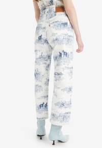 Pantalones de denim blanco con estampado de paisaje azul, diseño de pierna ancha y una etiqueta de marca de cuero en la cinturilla. Tejido texturizado con un patrón detallado.