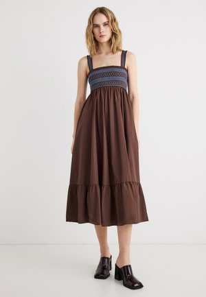 Γυναίκα που φοράει καφέ midi φόρεμα με μπλε σφηκωτή μπούστο και τιράντες, συνδυασμένο με μαύρα mule με τακούνι, στέκεται σε λευκό φόντο.