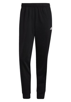 Man draagt zwarte trainingsbroek met drie witte strepen aan de zijkanten, witte sneakers en een wit shirt, staand met één been over het andere geslagen.