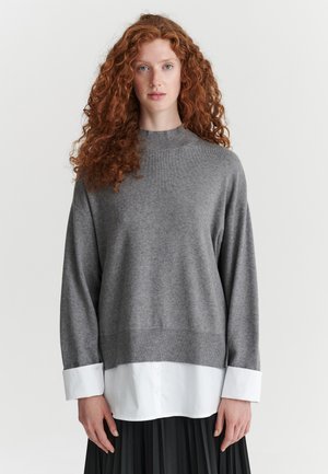 Femme aux longs cheveux roux bouclés portant un pull gris superposé à une chemise blanche et une jupe plissée noire, debout devant un fond uni.